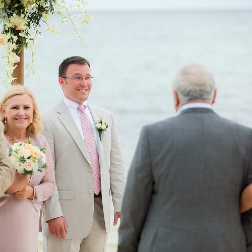 Groom watching bride walk down aisle on beach.