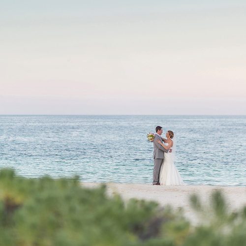Bride an groom on beach with green bush in foreground at Excellence Playa Mujeres