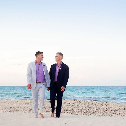 Grooms holding hands walking on beach in Cancun at wedding