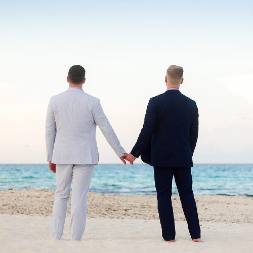 Grooms holding hands and looking out to sea at gay wedding in Cancun