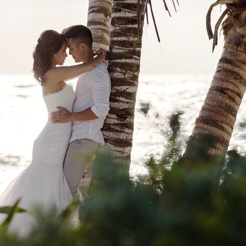 Bride and groom leaning up against a palm tree in romantic pose.