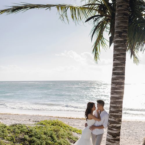 Bride and groom leaning up against a palm tree on beach in Riveira Maya Mexico