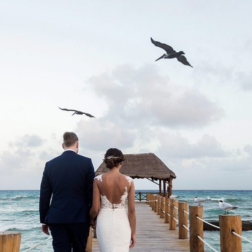 Bride and groom walking on dock with birds in the background in Riviera Maya