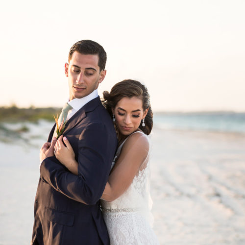 Bride hugging groom from behind on beach.