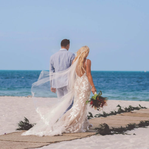 Bride and groom walking on beach with wind blowing veil.