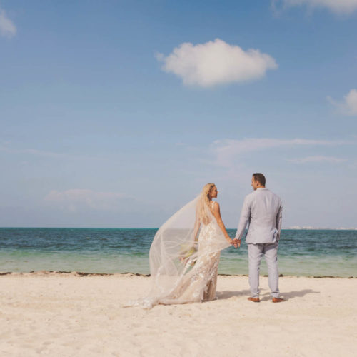 Wide angle photo of bride and groom holding hands and looking out at ocean with wind blowing veil.