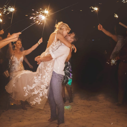 Groom lifting bride and kissing her under sparklers after wedding